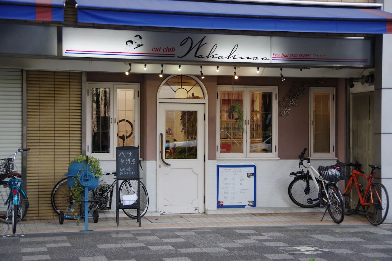 店內的白色雙門上掛著「cut club Wakana」的招牌，並提供捷徑和指甲花服務。 夾在窗戶和自行車停車場之間，入口附近有藍色的自行車架、室內植物、菜單和招牌。