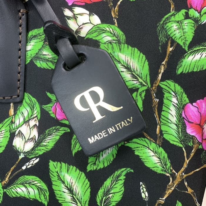 帶有金色“R”和意大利文字的黑色 ROBERTA PIERI 行李牌懸掛在帶有綠葉和粉紅色花朵的花卉面料上，與迷你行李袋相得益彰，具有永恆的意大利優雅。